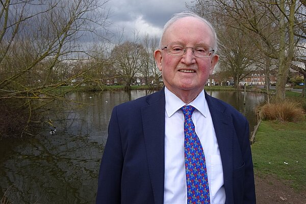 Clive Amos wearing jacket and tie in front of the pond in Theydon Bois